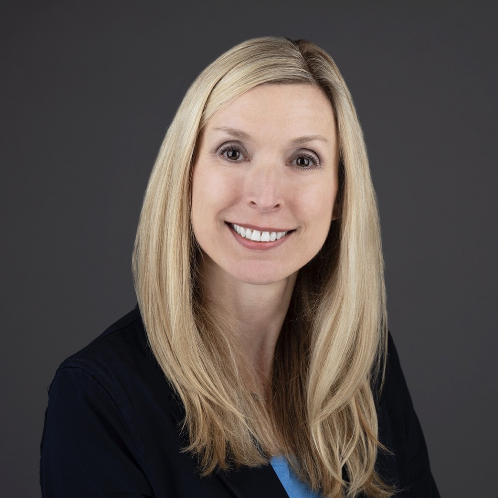 Professional headshot of a smiling woman with blonde hair wearing a black blazer against a gray background